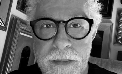Portrait of an older man with curly white hair, beard, and glasses, in a room with framed pictures on the wall.