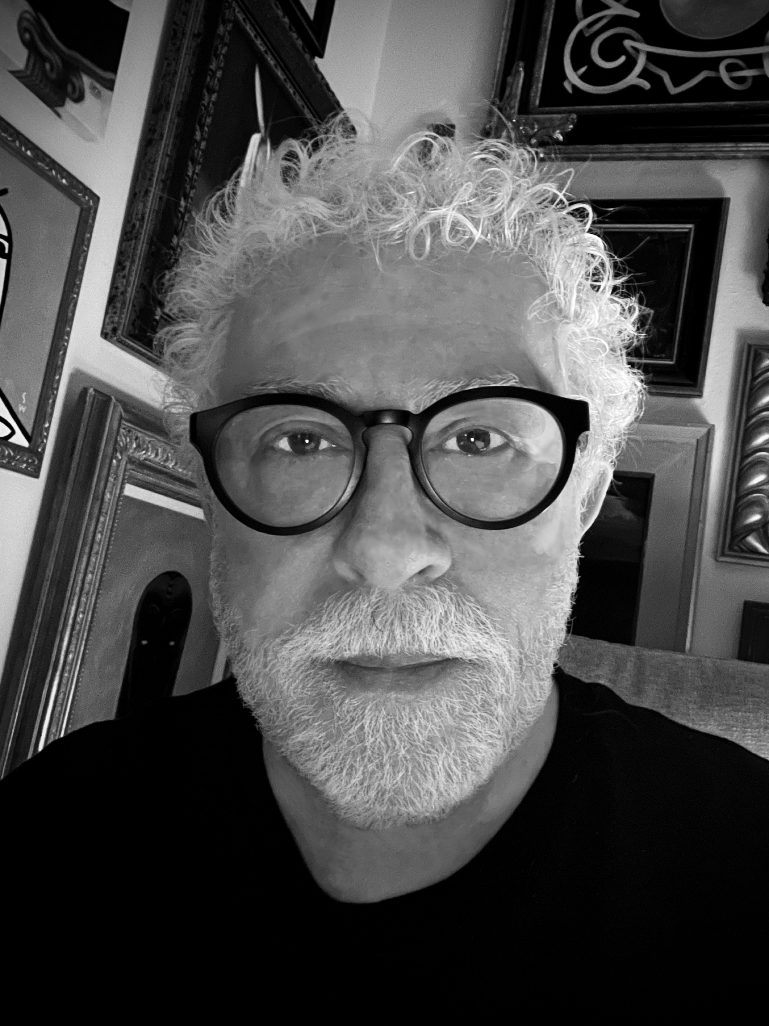 Portrait of an older man with curly white hair, beard, and glasses, in a room with framed pictures on the wall.