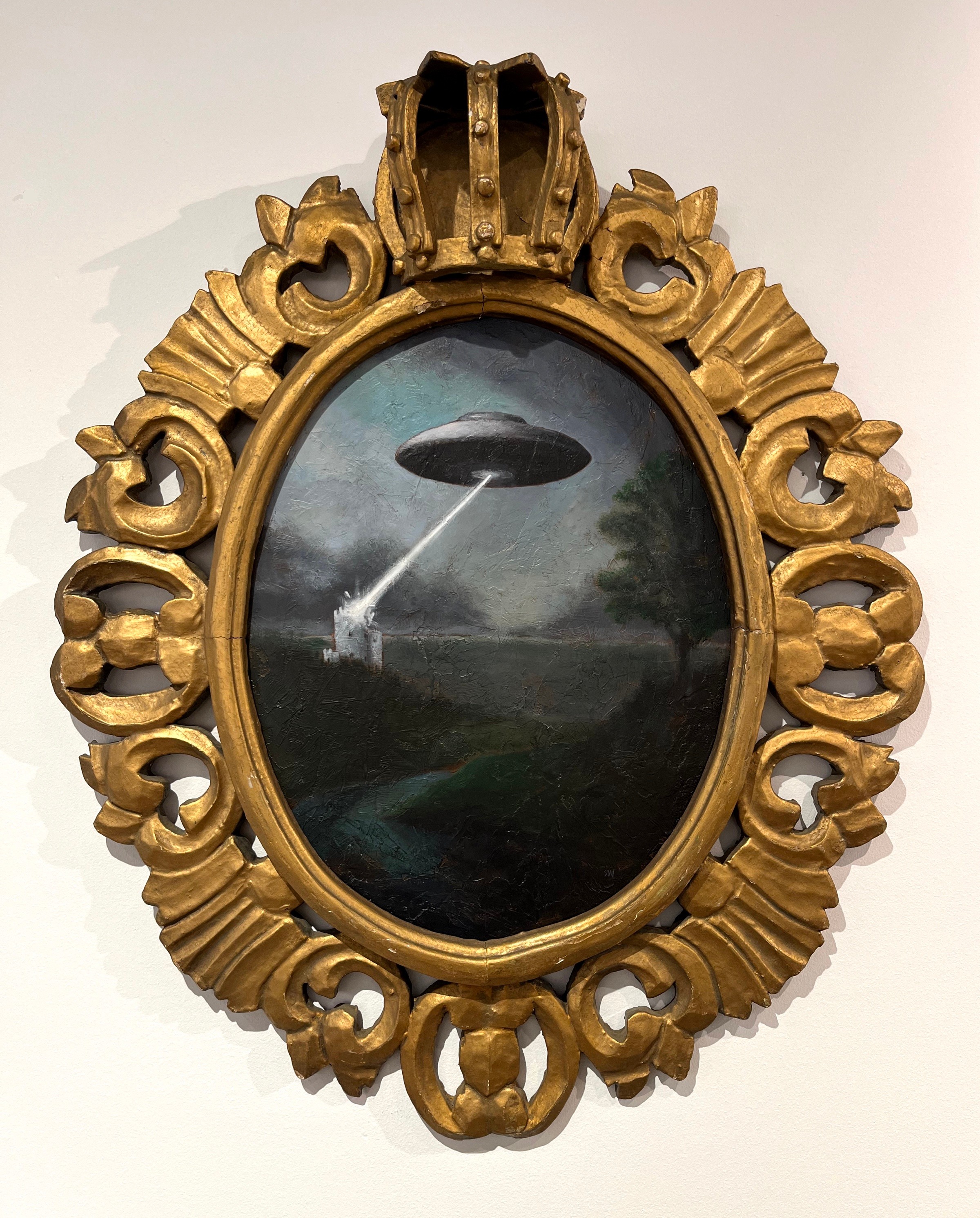 Oval framed painting depicts a UFO emitting a beam of light over a landscape with trees and a cloudy sky.