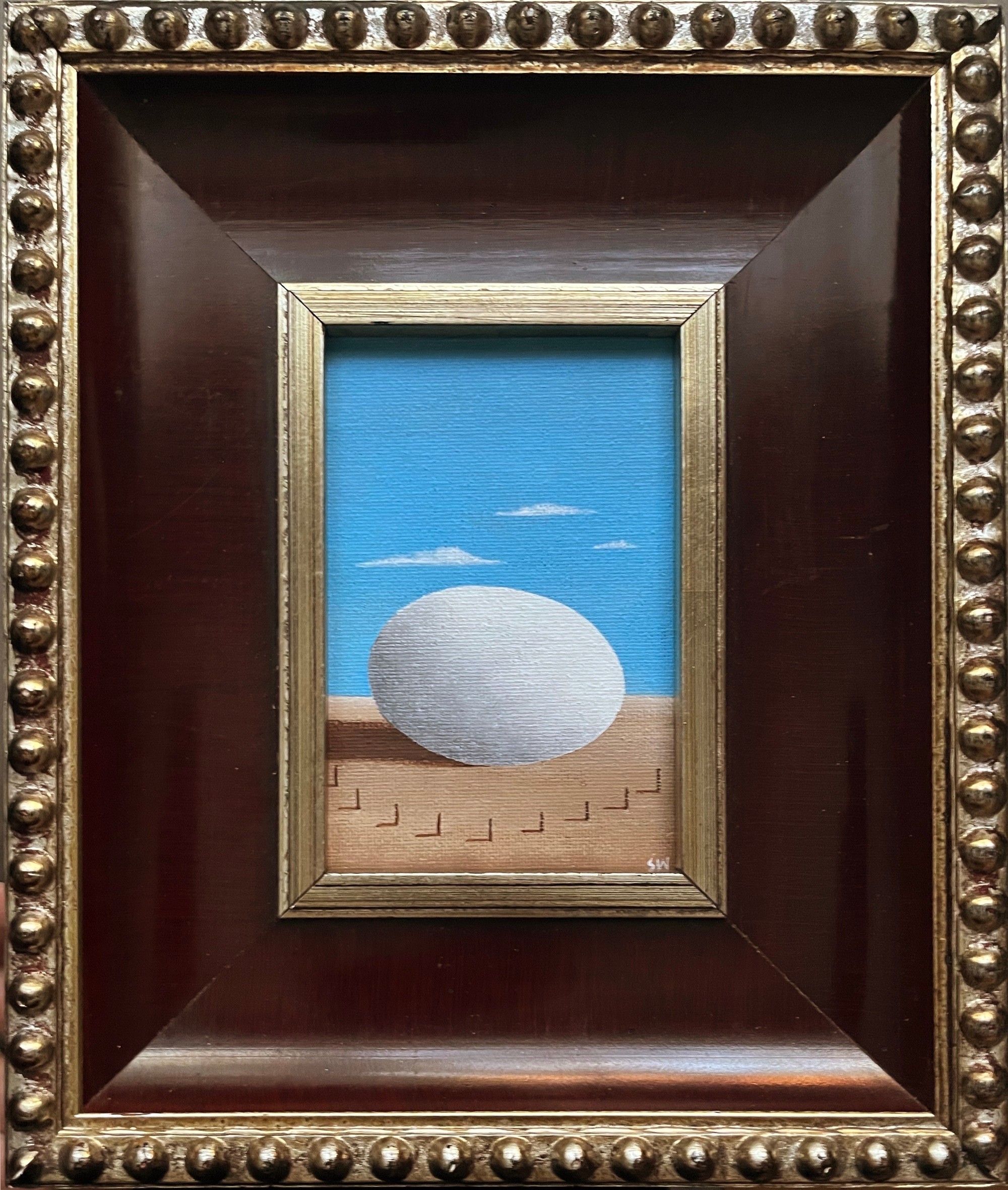 Small painting of a white egg on a sandy surface with a blue sky background, framed in a decorative wooden frame.