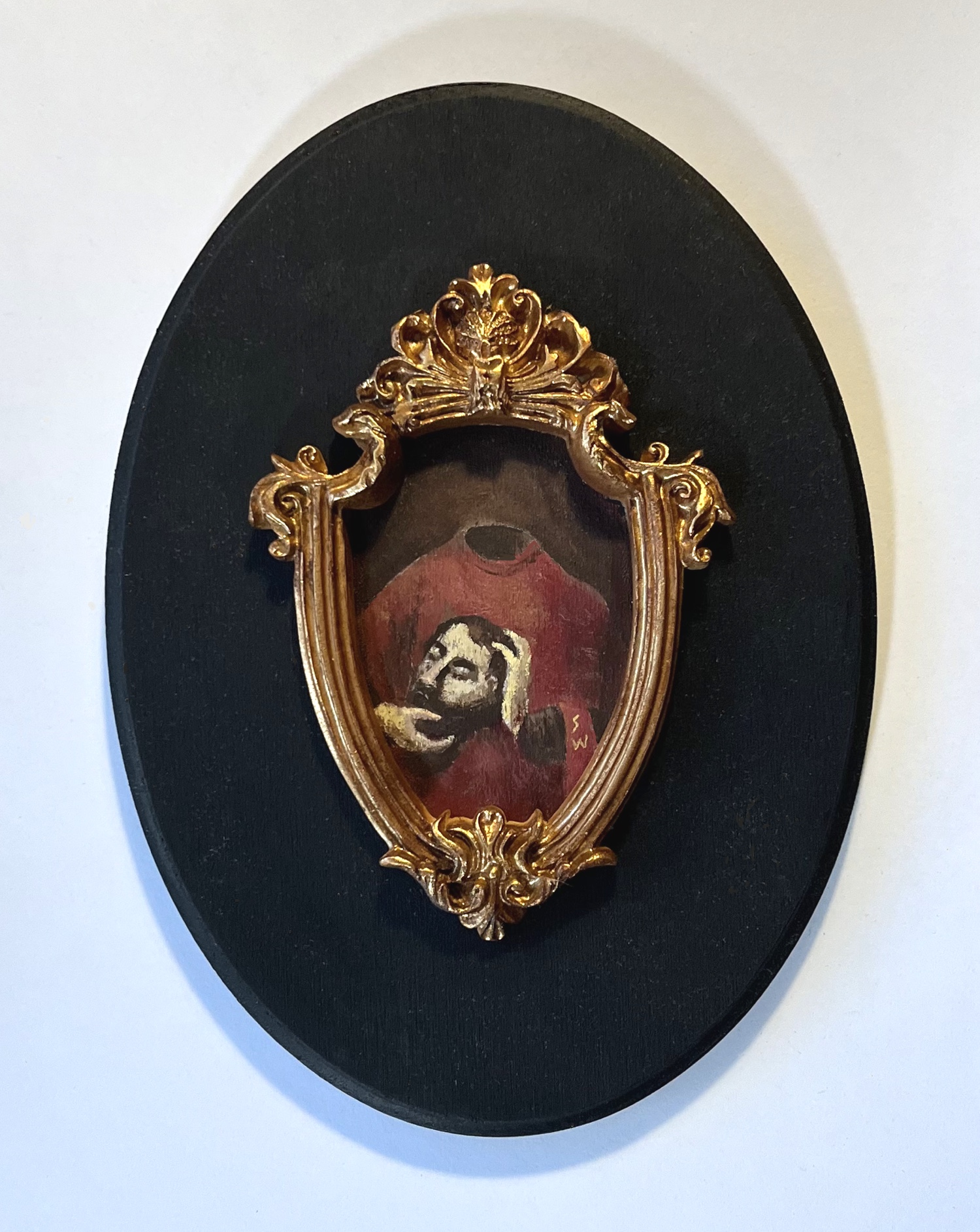 Small portrait of a person with a beard and hat, framed in an ornate gold frame, mounted on a black oval background.