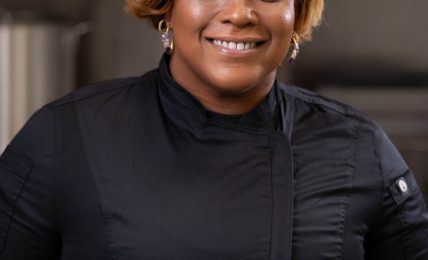 Smiling woman with short, wavy blonde hair wearing a black top, standing indoors.