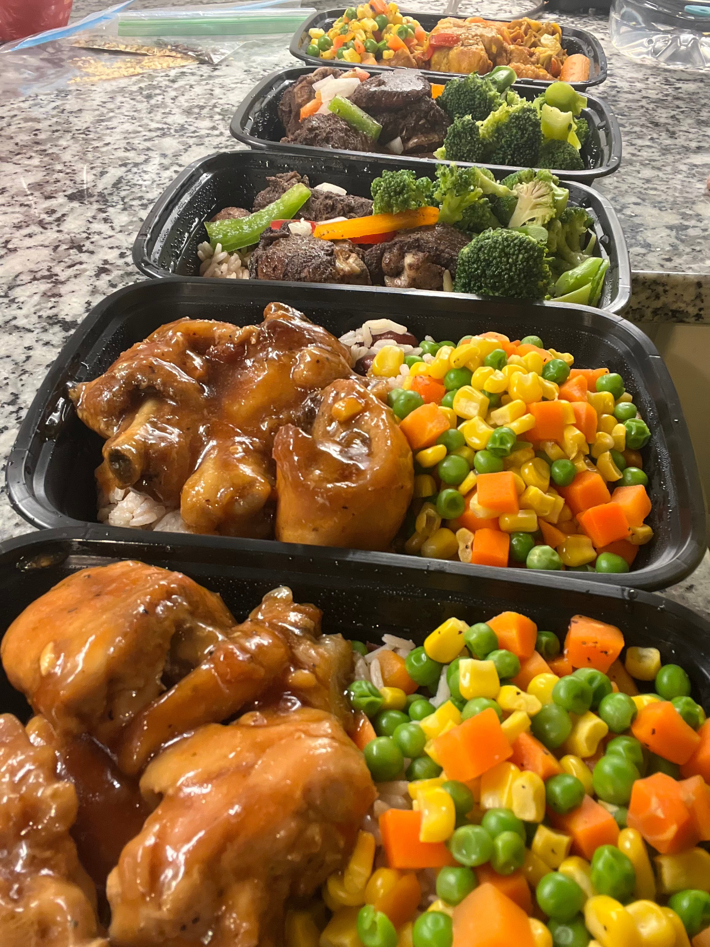 Four black meal containers filled with chicken, mixed vegetables, broccoli, carrots, peas, and corn, arranged on a granite countertop.