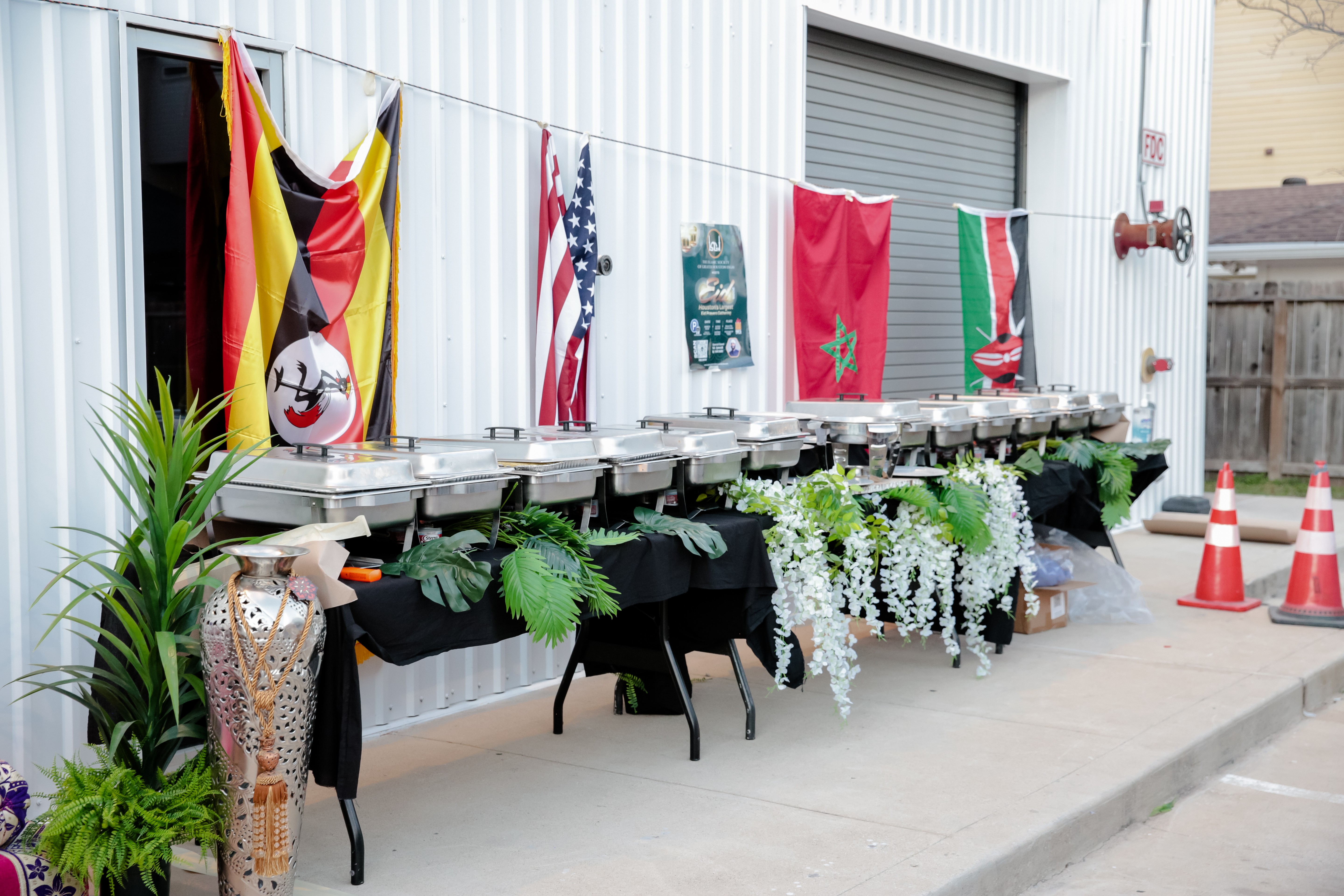 Clothesline with flags and decorations, table with plants and flowers, orange traffic cones, and a building with a garage door.