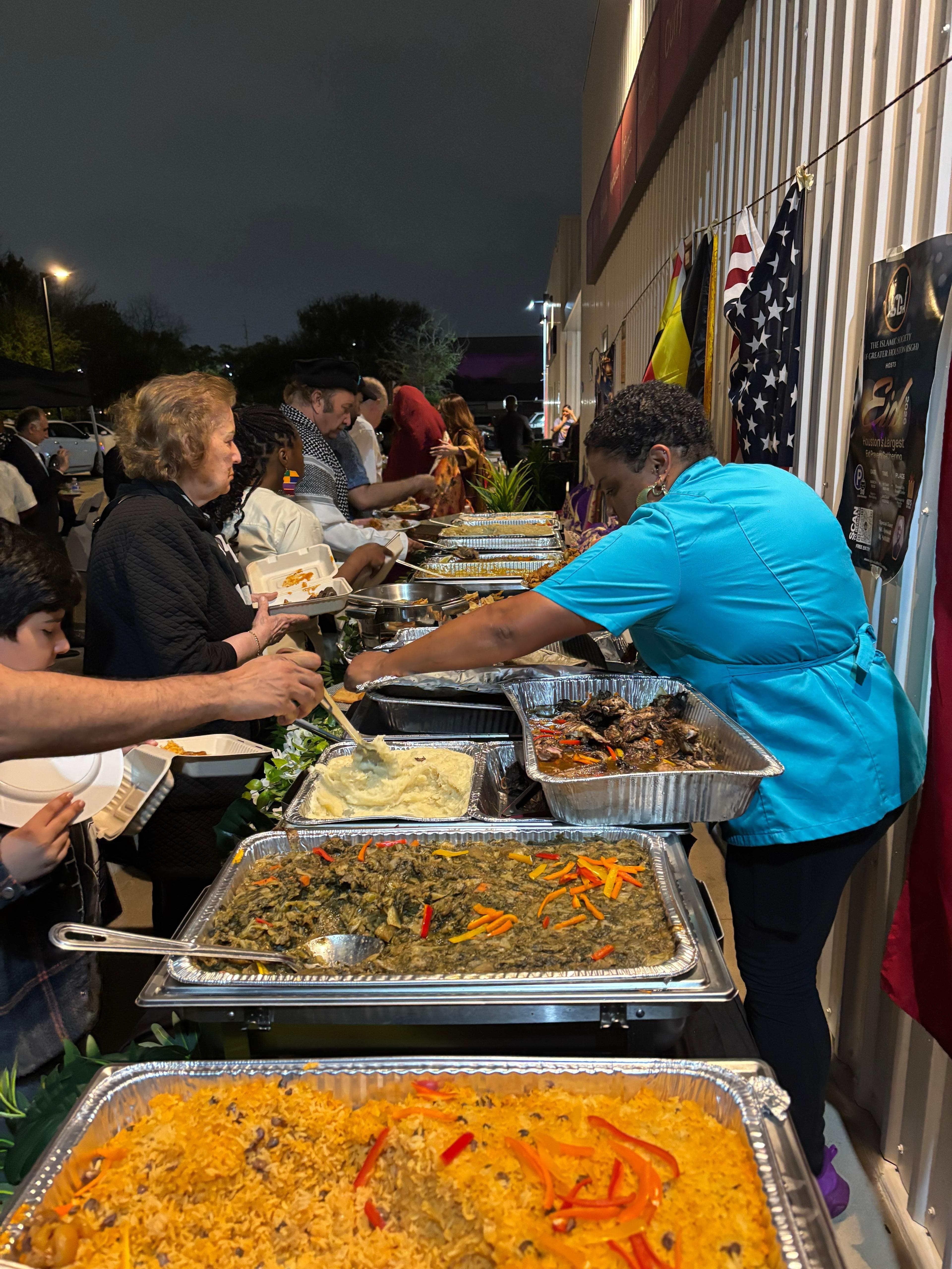 People line up at a food stand with trays of various dishes, including rice and vegetables, outdoors at night.