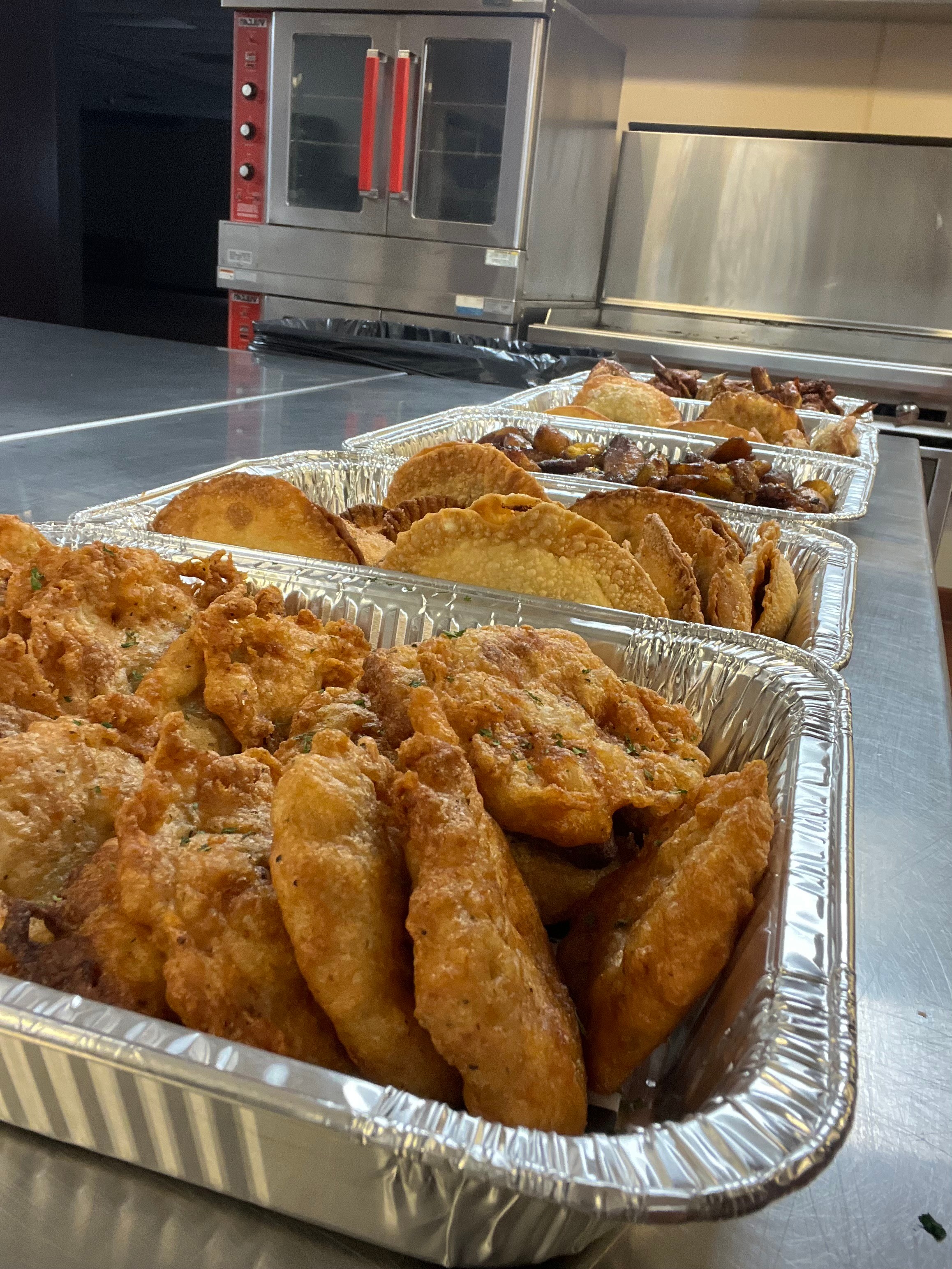 Multiple trays of fried chicken and other fried foods on a stainless steel counter in a kitchen.