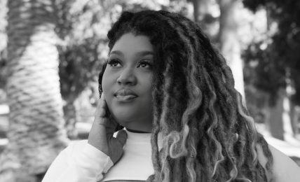 Young woman with long, curly hair outdoors, smiling, surrounded by trees, in black and white.
