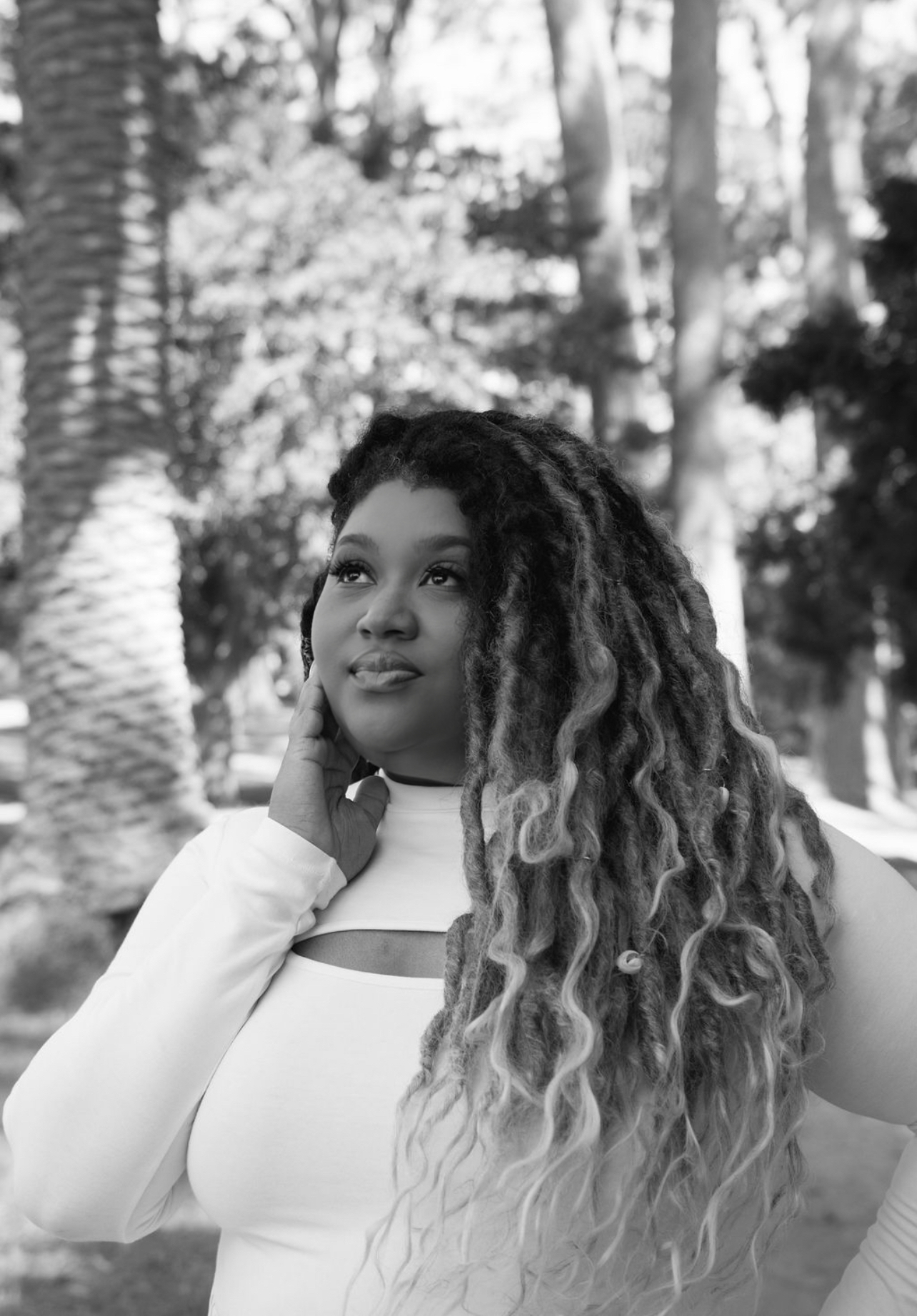 Young woman with long, curly hair outdoors, smiling, surrounded by trees, in black and white.