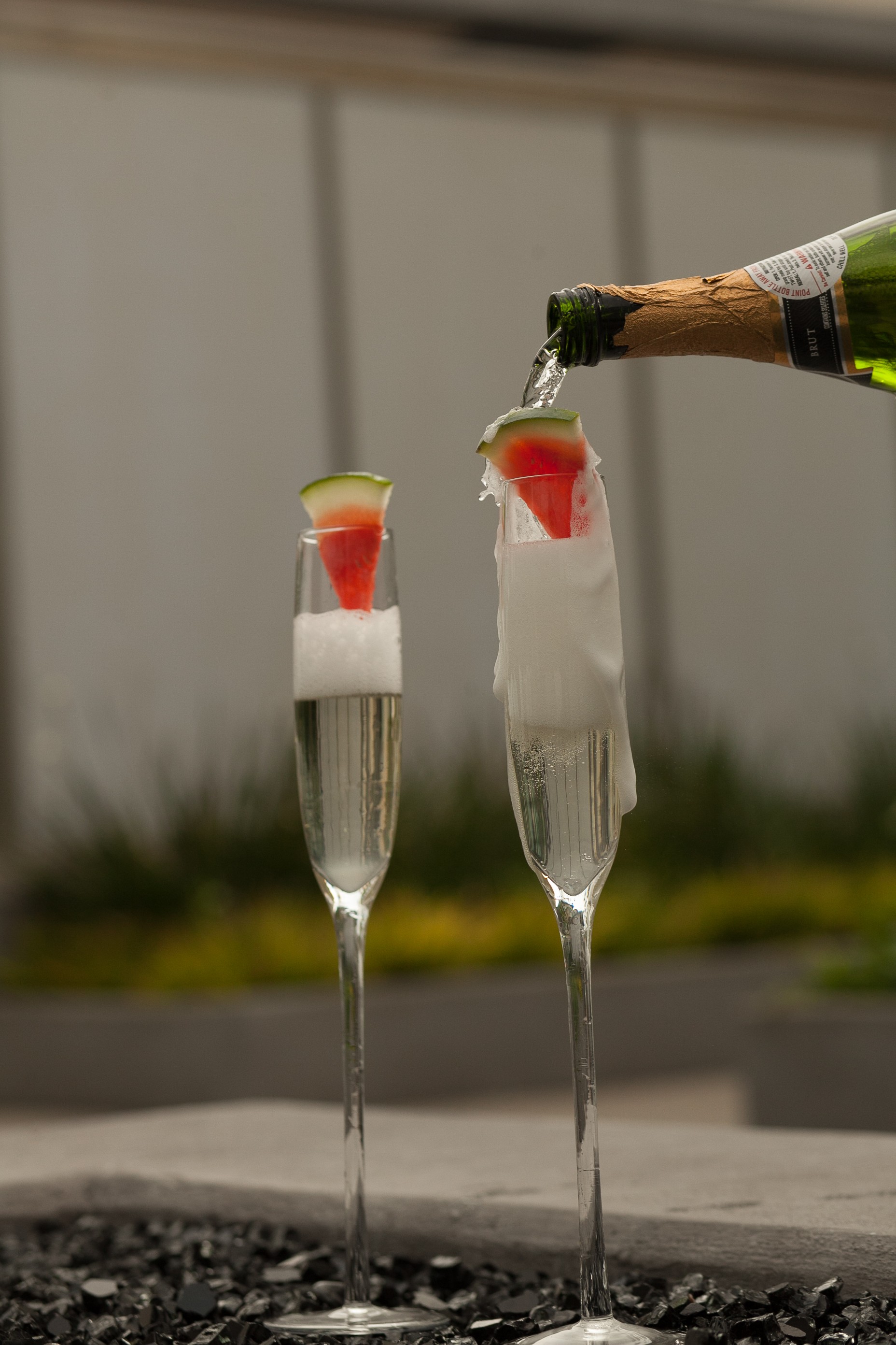 Two champagne flutes with garnishes, one being filled from a bottle, outdoors, with a blurred background.