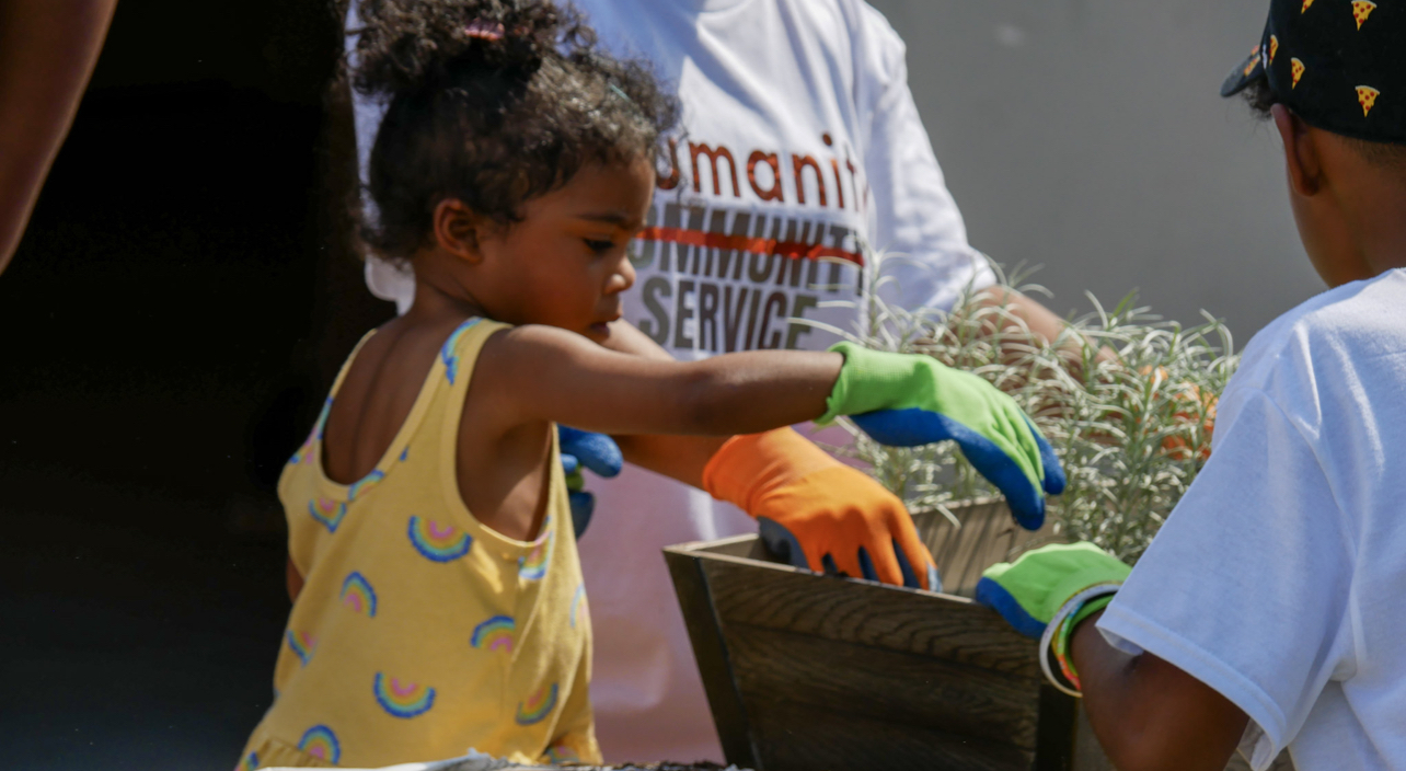 Child in yellow shirt receiving a tray from a person in gloves, outdoors, with plants nearby.