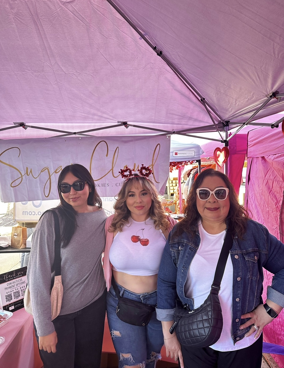 Three women stand under a pink canopy at an outdoor event, smiling and wearing sunglasses, with a sign behind them.
