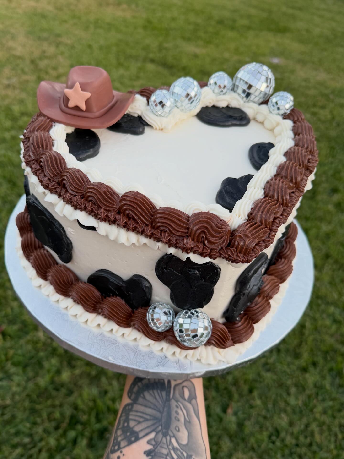 Heart-shaped cake decorated with chocolate, white, and black frosting, topped with a cowboy hat, silver disco balls, and black accents.