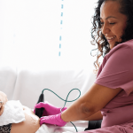 A woman in pink scrubs and pink gloves monitors a newborn baby in a hospital bed, smiling gently.