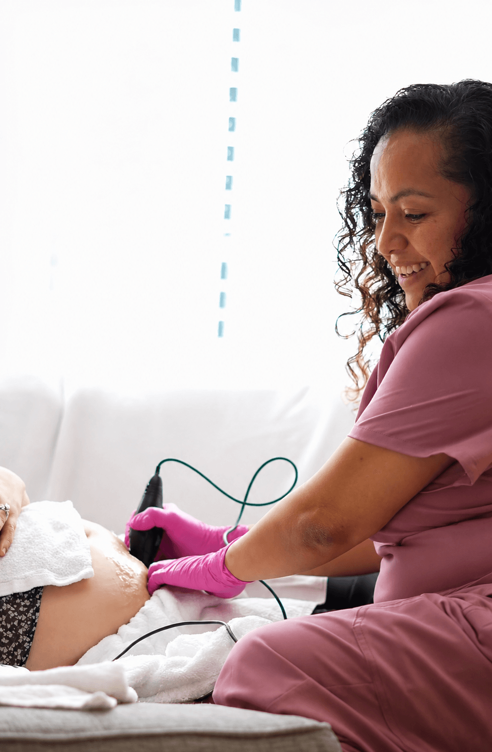 A woman in pink scrubs and pink gloves monitors a newborn baby in a hospital bed, smiling gently.
