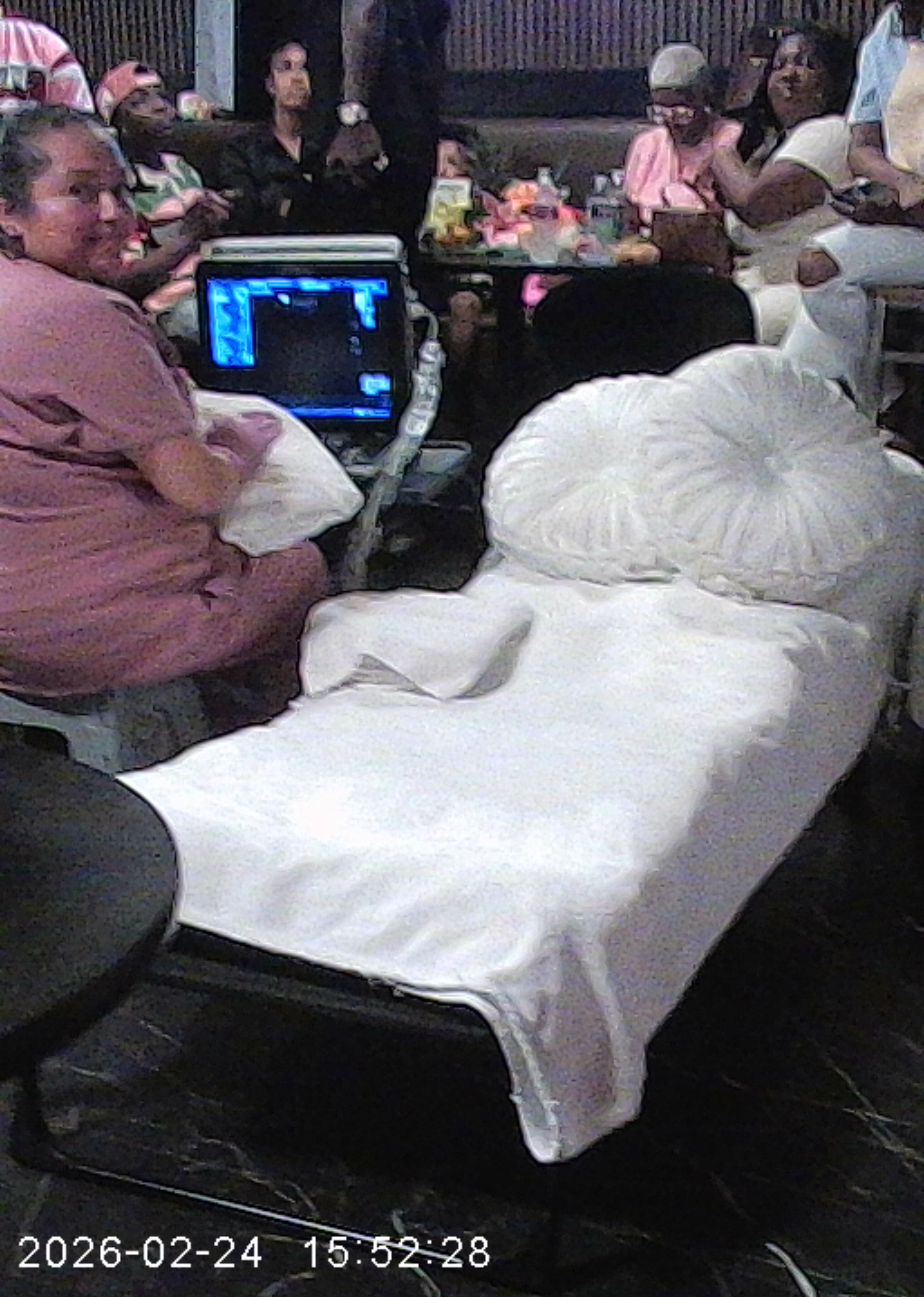 Hospital bed with white bedding and pillow, surrounded by people in a room, some using laptops.