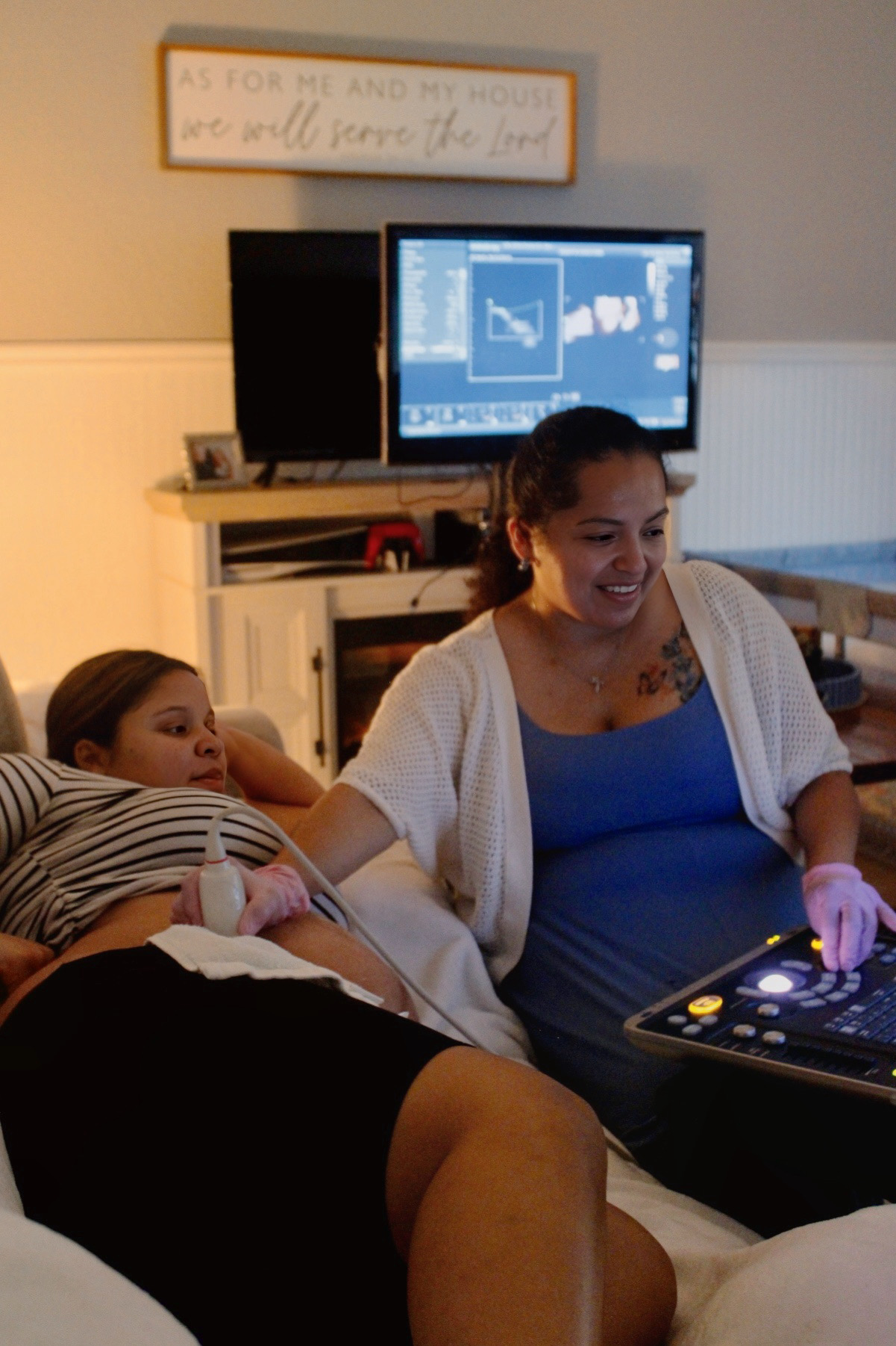Two women in a room, one sitting on a couch and the other using a medical device, with a computer screen in the background.