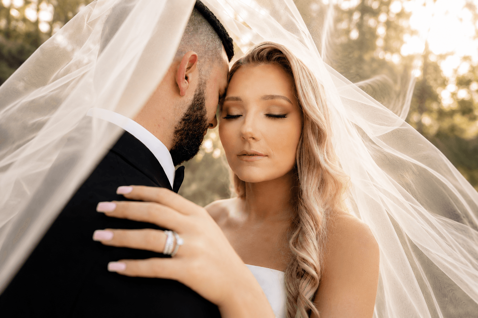Bride and groom under a veil, close-up of their faces and the bride's hand with a ring, outdoors with sunlight.