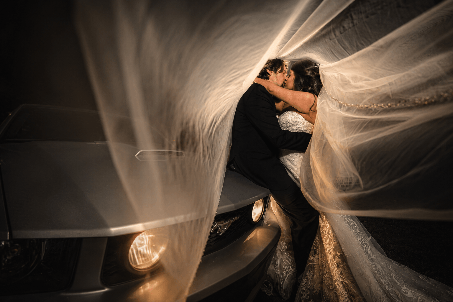 Bride and groom embrace under flowing wedding veil, with dark background and soft lighting, capturing an intimate moment.