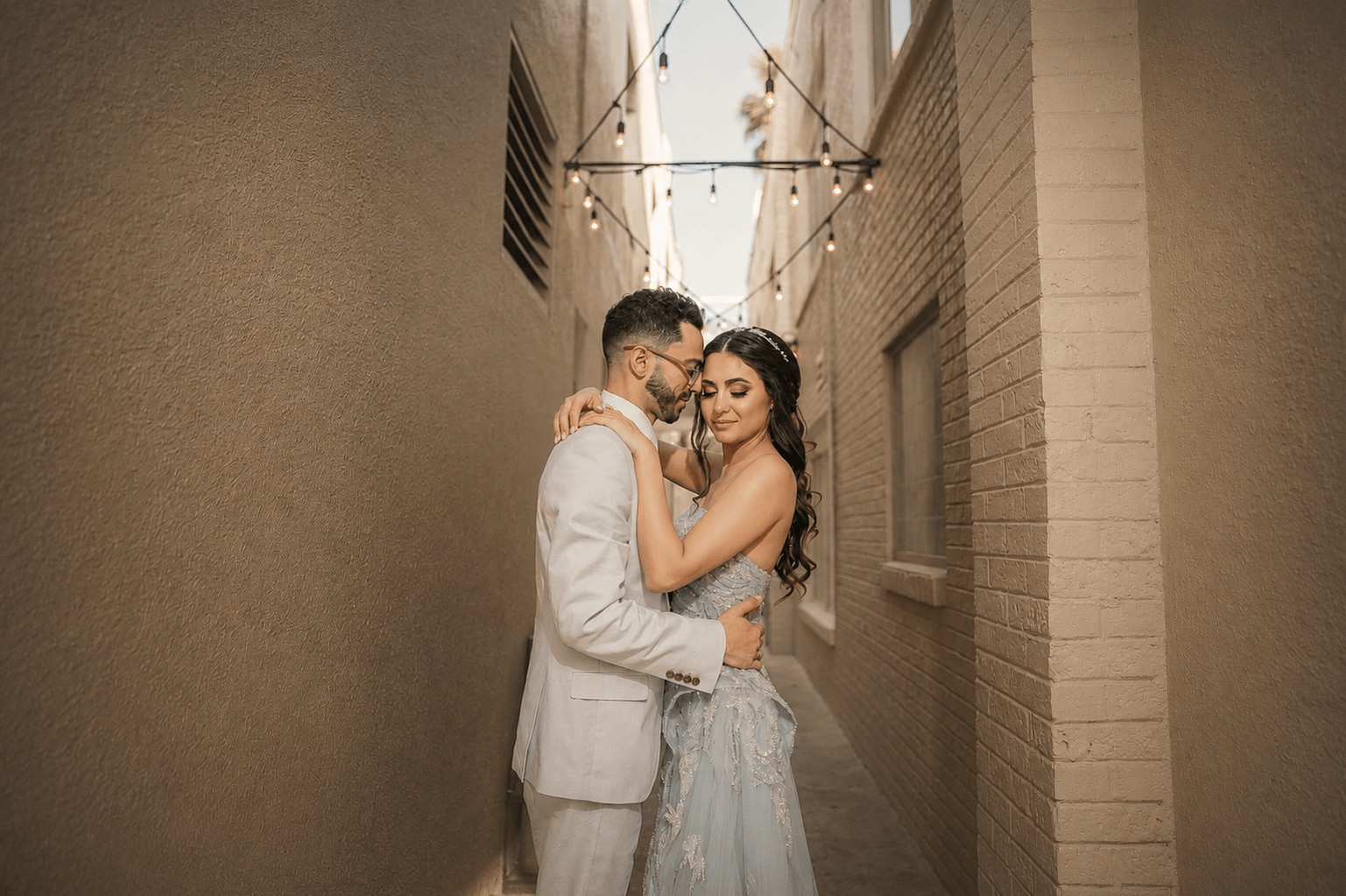 A couple embraces in a narrow alleyway with string lights overhead, the man in a light suit and the woman in a dress.