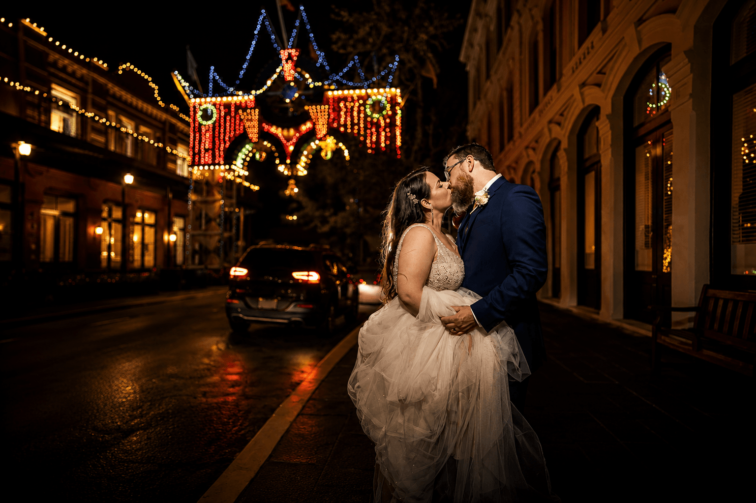 Couple in wedding attire kissing on a city street at night with festive holiday lights overhead.