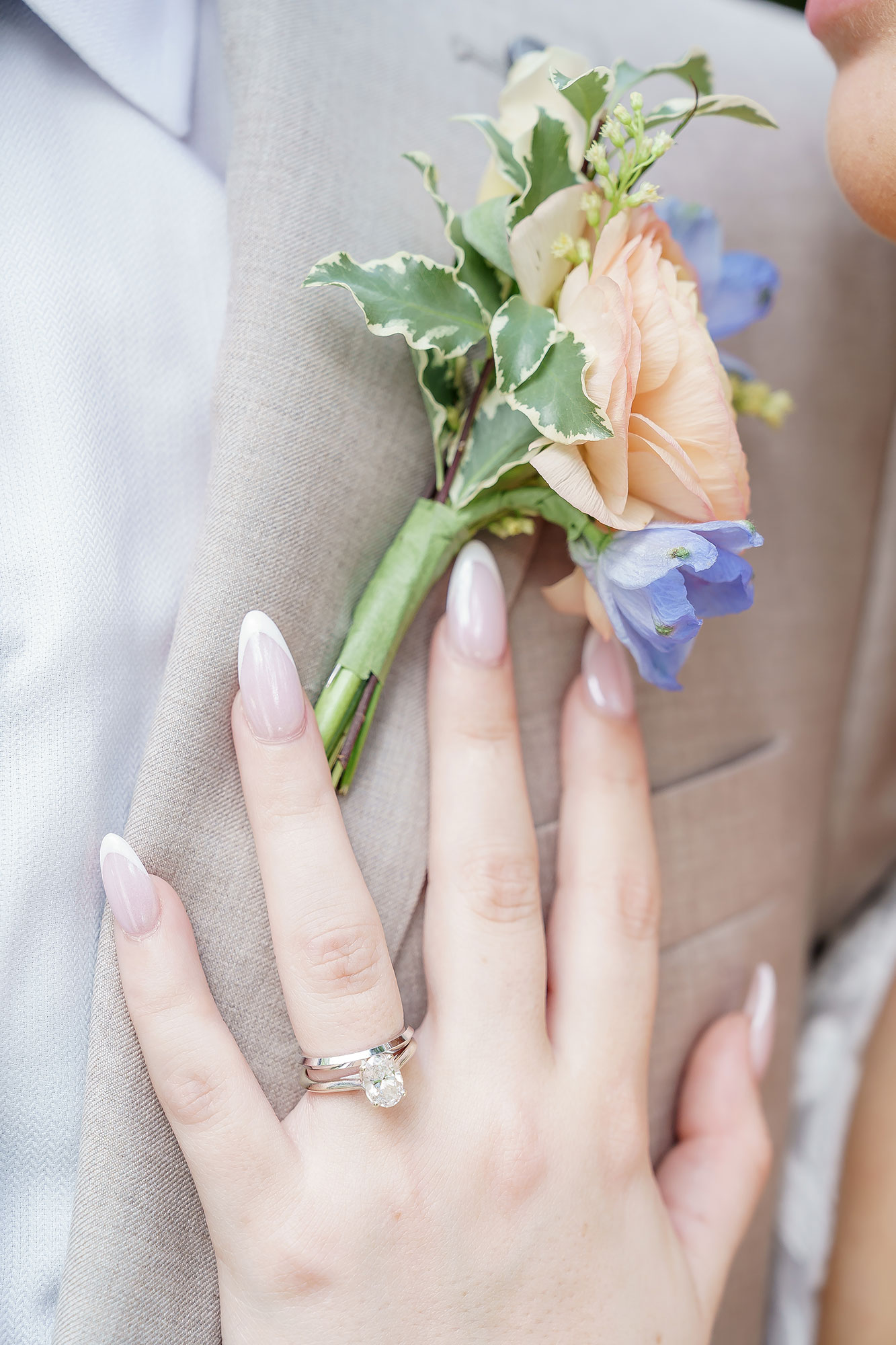 Person's hand with a ring holding a small bouquet of flowers, including a peach rose, blue flower, and green leaves.
