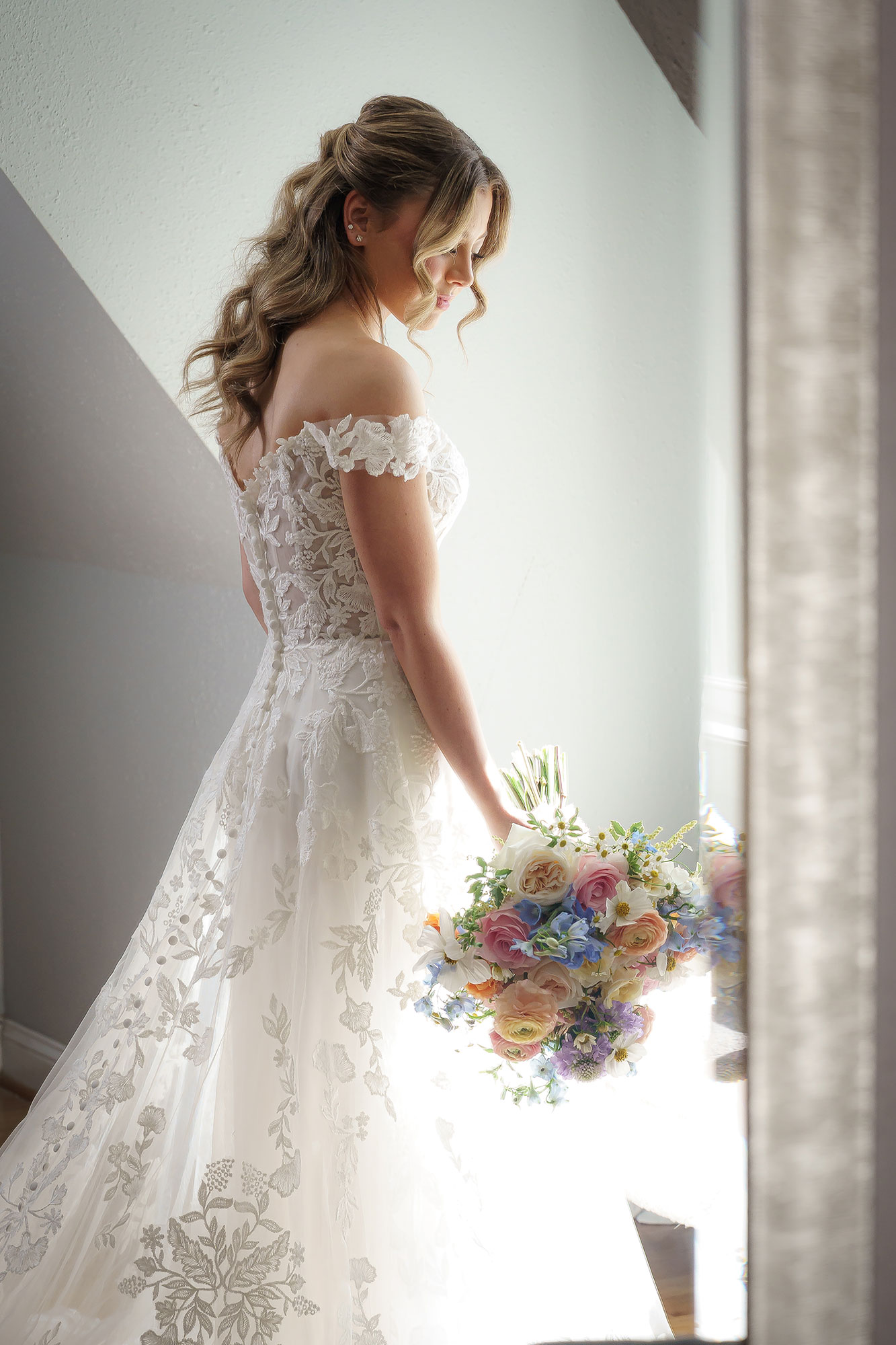 Woman in wedding dress holding a bouquet near a window, backlit, with light streaming in, side view.