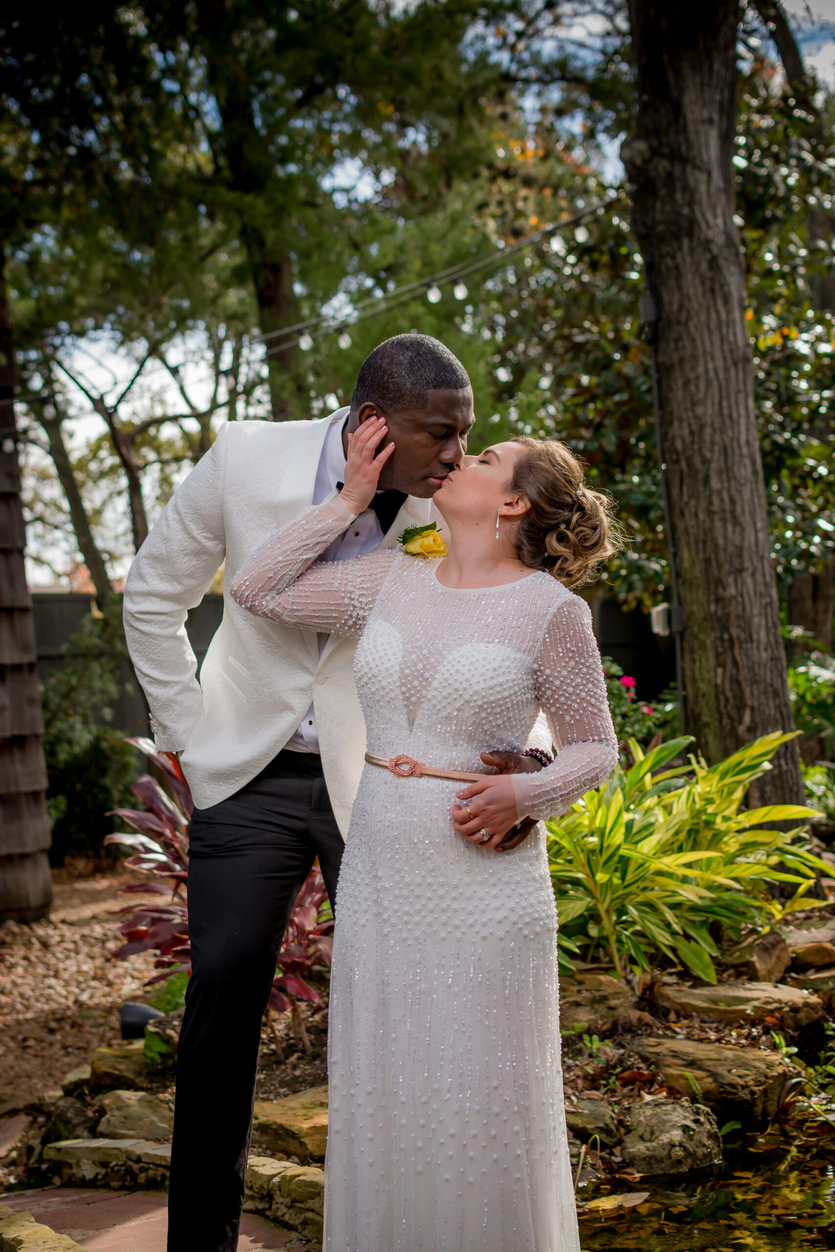 A couple stands outdoors among trees and plants, gazing at each other, with string lights overhead, in a garden setting.